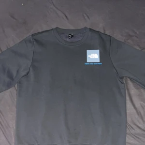 Svart tröja från The North Face - Säljer en stilren grå tröja från The North Face med deras logga tryckt på bröstet och ryggen. Tröjan har långa ärmar och en rund halsringning. Perfekt för en avslappnad stil.