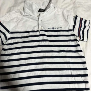 Randig t-shirt från Sail Racing - Säljer en snygg vit och marinblå randig t-shirt från Sail Racing. Den har korta ärmar och en klassisk krage med knappar. Perfekt för en avslappnad stil med en touch av segling.