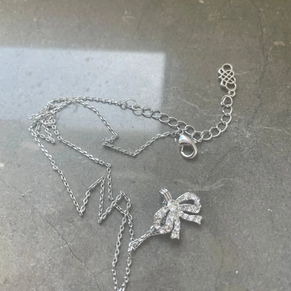 Elegant silverhalsband med en glittrig rosett som hänge. Kedjan är tunn och justerbar med ett klassiskt lås. Perfekt för att lägga till en gnistrande detalj till din outfit.. Asusteet.