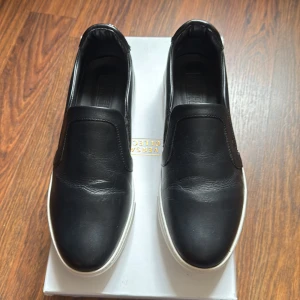 Svarta sneakers från Versace Collection - Jag säljer mina svarta sneakers från Versace Collection då jag bara använt dem 1-2 gånger och de har stått oanvända i garderoben. Skorna är i toppskick, med en stilren design, vit sula och rund tå i högkvalitativt skinn. Originalpriset var 7000 kr, men jag känner att jag inte använder dem tillräckligt för att behålla dem. Priset går att förhandla, så hör gärna av dig om du är intresserad!