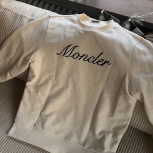 Vit sweatshirt från Moncler - Säljer en stilren vit sweatshirt från Moncler med deras logga broderad på bröstet. Tröjan har långa ärmar och en rund halsringning. Perfekt för en avslappnad och trendig look.