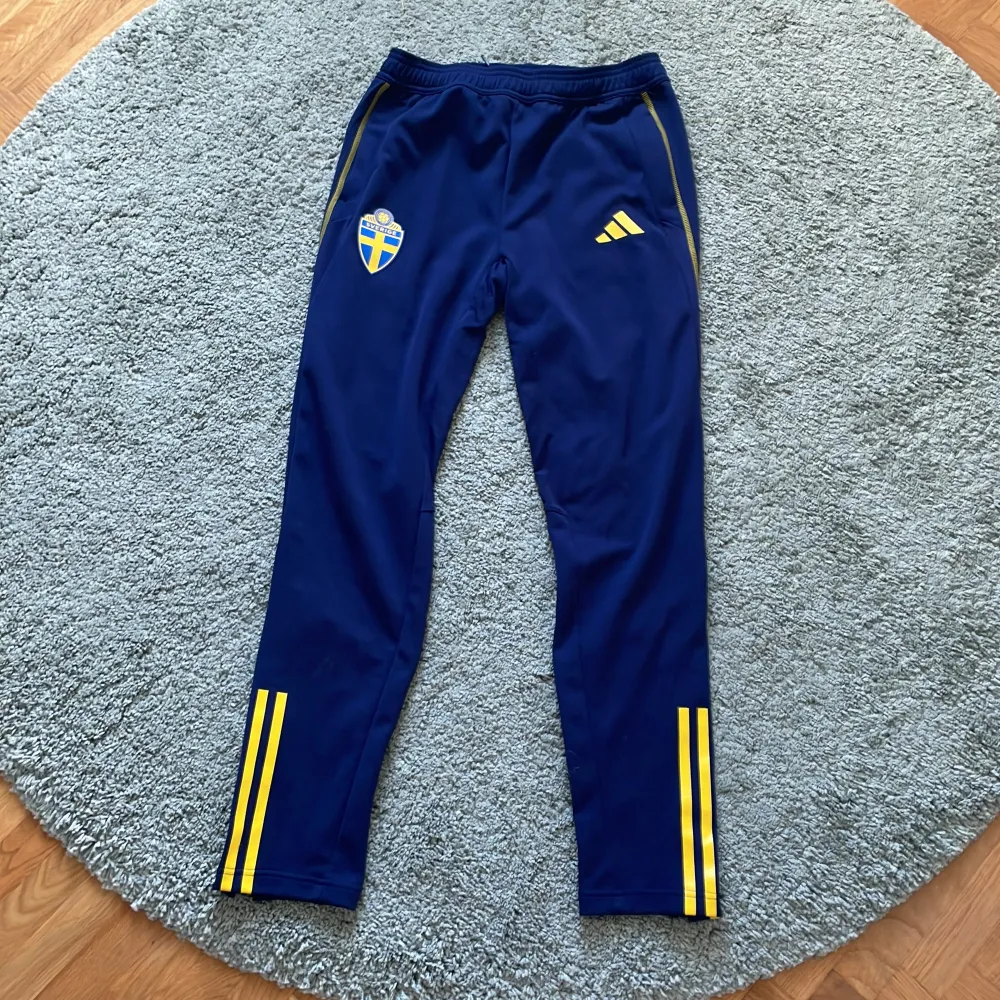 Snygga blå träningsbyxor från Adidas med Sveriges landslagslogga. Byxorna har gula detaljer och tre ränder längs benen. Perfekta för fotbollsträningen eller annan sportaktivitet. Elastisk midja för bekväm passform. Sköna och knappt använda. Hör av er vid fundering!. Farkut & Housut.