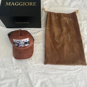 Brun keps från Maggiore - Snygg brun keps från Maggiore med ett svartvitt tryck framtill. Kepsen har ett nätmaterial på baksidan och justerbar passform. Perfekt för en avslappnad stil. Modell C4