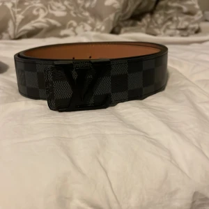 Rutig bälte med monogramspänne - Snyggt bälte med ett rutigt mönster i svart och grått. Bältet har ett monogramspänne som ger en lyxig känsla. Perfekt för att lyfta vilken outfit som helst.