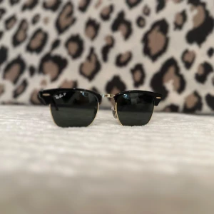 Svarta solglasögon från Ray-Ban - Snygga svarta solglasögon från Ray-Ban med gulddetaljer. De har en klassisk design med mörka glas och en stilren look. Perfekta för soliga dagar och kommer med ett svart fodral för säker förvaring.