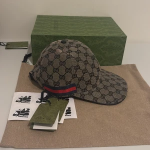  keps från Gucci Canvas Navy - Snygg keps från Gucci med det ikoniska GG-mönstret i beige och svart. Kepsen har en justerbar rem baktill och en röd och blå randdetalj på sidan. Perfekt för att ge din outfit en lyxig touch. Finns i S M L