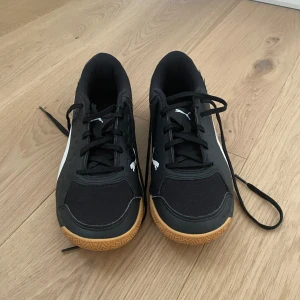 Svarta sneakers från Puma - Snygga svarta sneakers från Puma med vit logga och detaljer. Skorna har en rund tå och snörning framtill. Sulan är i en kontrasterande gummifärg som ger ett sportigt intryck. Perfekta för träning inomhus och bollspel.