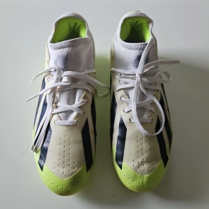 Adidas fotbollsskor med neongula detaljer - Snygga Adidas fotbollsskor i vitt med neongula och svarta detaljer. Skorna har snörning och en modern design med tydliga ränder. Perfekta för fotbollsplanen med en greppvänlig sula. Använda en säsong och endast på gräs. 