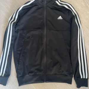 Svart träningsjacka från Adidas - Säljer en klassisk svart träningsjacka från Adidas med de ikoniska tre ränderna längs ärmarna. Jackan har dragkedja framtill och ribbade muddar vid ärmslut och nederkant. Perfekt för träning eller en sportig look. Strl 164. Rök och djurfritt hem.