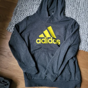 Grå hoodie från Adidas - Säljer en snygg grå hoodie från Adidas i storlek 152/158. Tröjan har en klassisk känguruficka och en bekväm huva. Använd en del men inga defekter. Priset är absolut förhandlingsbart, så tveka inte på att höra av dig.