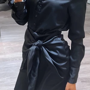Svart långärmad klänning med knyt - Elegant svart klänning i satin med långa ärmar och knyt i midjan. Perfekt för en stilren look. Storlek S från BikBok 