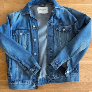 Blå jeansjacka från These Glory Days - Säljer en klassisk blå jeansjacka från These Glory Days. Jackan har en normal passform med långa ärmar och knappar framtill. Två bröstfickor med knappstängning ger en stilren look! Se bild 4 för en liten fläck på underarmen annars Cond 9/10