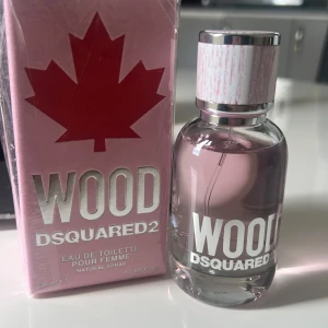 Wood Eau de Toilette från DSQUARED2 - Säljer en elegant Eau de Toilette från DSQUARED2 med en fräsch och träig doft. Flaskan är genomskinlig med rosa vätska och har en stilren design med ett lönnlöv på korken. Förpackningen är rosa med ett stort rött lönnlöv. Perfekt för den som älskar en naturlig och sofistikerad doft.