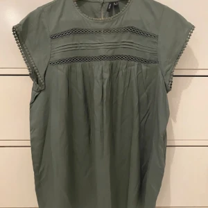 Grön blus från Vero Moda - Säljer en stilren grön blus från Vero Moda. Blusen har korta ärmar och dekorativa spetsdetaljer vid ärmslut och över bröstet. Perfekt för en avslappnad men ändå elegant look. 💚