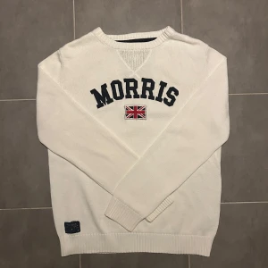 Morris Stickad Tröja Creme L - Säljer en stilren vit tröja från Morris i storlek L. Tröjan är tillverkad i 100% bomull och har ett tryck med texten 'Morris' och en brittisk flagga på framsidan. Perfekt för en avslappnad och snygg look.