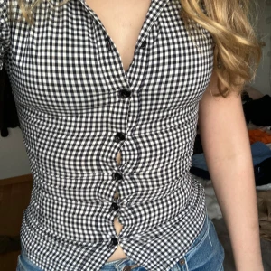 Svartvit rutig blus - Snygg svartvit rutig blus med knappar framtill. Blusen har en figurnära passform och korta ärmar, perfekt för en stilren look. Passar bra till både jeans och kjol.