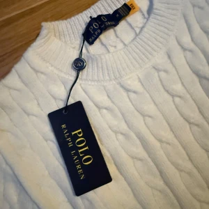 Vit tröja Polo Ralph Lauren, helt ny | stickad - Säljer en stilren vit stickad tröja från Polo Ralph Lauren med klassiskt kabelstickat mönster. Tröjan har långa ärmar och en rund halsringning. Perfekt för en elegant och avslappnad look.
