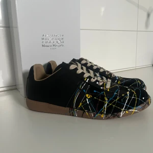 Maison Margiela Gats - Unika svarta sneakers från Maison Margiela med ett färgstarkt stänkmönster i gult, blått och vitt. Skorna har en rund tå och snörning, vilket ger en klassisk men ändå modern look. Perfekta för dig som vill sticka ut med stil. De är använda ett par gånger och skicket är 9/10. Man får med dustbags och låda. Passar någon smått. Vid frågor så är det bara att skriva!