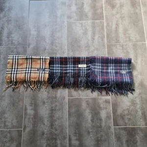 Rutiga halsdukar från Burberry - Säljer två den i mitten är såld snygga rutiga halsdukar från Burberry i olika färger. Den första är beige med klassiskt rutmönster, den andra såld !!!är svart med blå och röda rutor, och den tredje är blå med röda och grå rutor. Perfekta för att hålla dig var