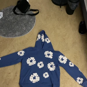 Blå hoodie med blommönster från Denim Tears - Säljer en blå hoodie från Denim Tears med ett unikt vitt blommönster. Hoodien har en klassisk design med långa ärmar och en bekväm huva. Perfekt för en avslappnad stil. Passar mer L