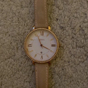 Beige klocka från Fossil - Elegant klocka från Fossil med en beige skinnrem och en glittrig boett i guld. Urtavlan är vit med romerska siffror och små kristaller som markörer. Perfekt accessoar för att lyfta din stil.