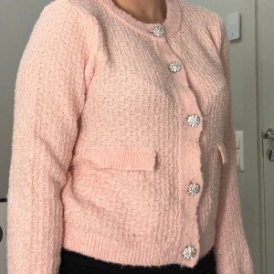 Rosa stickad kofta med glittriga knappar - Säljer en söt rosa stickad kofta med glittriga knappar. Koftan har en rund halsringning och långa ärmar. Perfekt för att lägga till lite glamour i vardagen. Kontakta mig för mer info! 💖