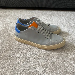 Axel Arigato Triple Clean 90 Sneakers - Snygga grå sneakers från Axel Arigato med blå häl och orange plös. Skorna har en rund tå och snörning framtill. Perfekta för en stilren look.