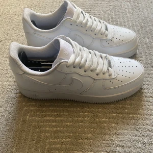 Vita Nike Air Force sneakers - Säljer ett par klassiska vita Nike Air Force sneakers. De har en stilren design med snörning och perforerade detaljer på tån för extra ventilation. Perfekta för en casual look.