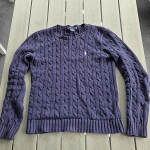 Mörkblå stickad tröja från Ralph Lauren - Säljer en snygg mörkblå stickad tröja från Ralph Lauren med kabelstickat mönster och broderad logga på bröstet. Tröjan har långa ärmar och ribbstickade muddar vid ärmslut och nederkant. Tröjan är i top skick då den har använts 2 gånger och det finns ibga defekter 