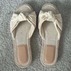 Beige sandaler från H&M - Säljer ett par stilrena beige sandaler från H&M. De har en knutdetalj på ovansidan och är tillverkade i ett linneliknande material. Perfekta för varma sommardagar! Helt oanvända