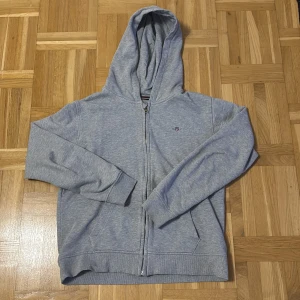 Grå hoodie från GANT - Säljer en stilren grå hoodie från GANT med dragkedja och broderad logga på bröstet. Perfekt för en avslappnad look. Den har en klassisk passform och är tillverkad i mjukt material för extra komfort.