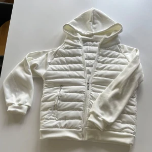 Vit cardigan jacka med huva - Snygg vit jacka med huva och dragkedja framtill. Jackan har quiltade partier på bröst och rygg, släta ärmar och två fickor med dragkedja. Perfekt för dig som gillar en clean och sportig look. Materialet känns mjukt och lätt.