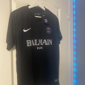 Svart PSG x Balmain fotbollströja Nike - Svart fotbollströja från Nike i samarbete med PSG och Balmain. Tröjan har korta ärmar, rund hals och är tillverkad i Dri-FIT-material som andas. Stora vita tryck med Balmain Paris och PSG-logga på bröstet. Perfekt för dig som gillar stilren och exklusiv sportstil.