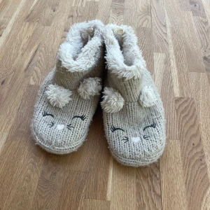 Beige stickade tofflor med kattansikte H&M - Supermysiga beige stickade tofflor från H&M med gulligt broderat kattansikte och fluffiga öron. Perfekta för att hålla fötterna varma hemma. Mjukt och fluffigt material både in- och utvändigt.