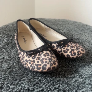 Leopardmönstrade ballerinaskor med rosett - Snygga ballerinaskor från Nobaci med leopardmönster i beige och svart. Skorna har rund tå, svarta kanter och en liten rosett framtill. Perfekta för dig som vill sticka ut med ett djurmönster och ändå hålla det klassiskt.