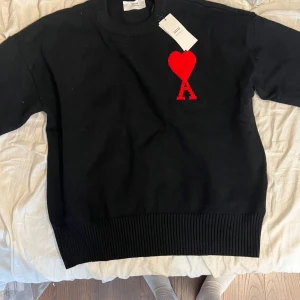 Svart stickad tröja från Ami Paris - Svart stickad tröja från Ami Paris med rund halsringning och ribbade muddar. På bröstet finns ett rött hjärta med bokstaven A, vilket ger en ikonisk look. Perfekt för dig som gillar stilrena plagg med en twist. Sitter som storlek M