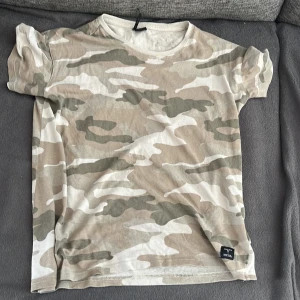 Beige camo t-shirt från 157 - Snygg t-shirt från 157 med beige, grå och vita camouflage-mönster. Klassisk rund halsringning och korta ärmar. Perfekt för dig som gillar streetstyle och vill sticka ut lite extra. Mjuk och skön bomullskänsla.