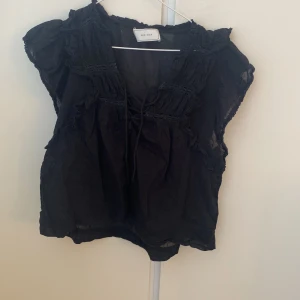 Svart blus med volang  - Supersöt svart blus från Nelly i storlek 36 i luftig bomull. Blusen har volangdetaljer vid axlar och hals, knytning framtill och är ärmlös. Snygg till både jeans och kjol! Säljer då den inte kommer till användning💗