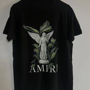 Svart Amiri t-shirt med ängeltryck - Svart t-shirt från Amiri med coolt tryck av en ängel och gröna blad på ryggen samt Amiri-logga. Framsidan har Amiri-tryck med färgstänk som rinner ner. Klassisk passform och rund hals. Perfekt för dig som gillar streetwear och snygga detaljer.