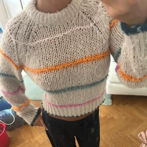 Beige stickad tröja med färgglada ränder - Supermysig beige stickad tröja med breda, färgglada ränder i orange, blått, rosa och vitt. Tröjan har en croppad passform, lång ärm och ribbad kant vid hals, ärmslut och nederkant. Perfekt för dig som gillar en chill och färgglad stil.