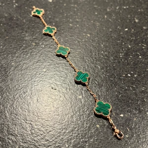 Grönt fyrklöver armband i guldton - Superfint armband med fem gröna fyrklöverformade hängen och guldfärgad kedja. Hängena har en snygg randig effekt i stenen och detaljerad kant. Perfekt accessoar för att lyfta din stil med en färgklick och cool design.