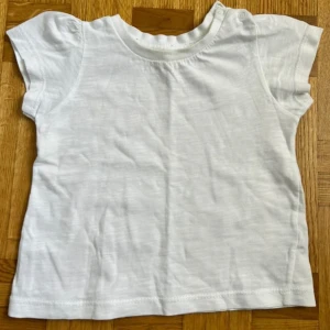 Vit t-shirt från Hello Baby - Enkel och mjuk vit t-shirt från Hello Baby i bomull. Modellen har korta ärmar, rund halsringning och tryckknappar på axeln för enkel påklädning. Perfekt basic-plagg till sommaren.