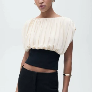 Svart och vit croppad blus med öppen rygg - Säljer en unik croppad blus i svart och vit med puffig, plisserad överdel och tight midja. Toppen har en snygg öppen rygg och är ärmlös med bred båturringning. Perfekt för dig som vill sticka ut med en cool och elegant look. Nyskick med prislapp. 