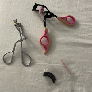 Två ögonfransböjare + extra gummidynor - Säljer två ögonfransböjare, en i klassisk silverfärgad metall och en med svart och beige handtag med rosa detaljer. Medföljer två extra svarta gummidynor i plastpåse. Perfekt för att få snyggt böjda fransar.