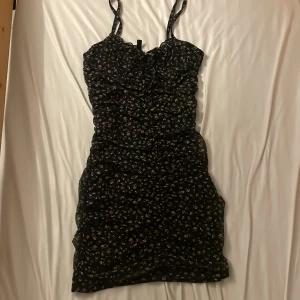 Svart blommig kortklänning H&M S - Supersöt svart kortklänning från H&M med små blommor i vitt och ljusrosa. Klänningen har smala axelband, rynkade detaljer och en liten rosett framtill. Perfekt för dig som gillar en feminin och trendig look. Perfekt till sommar! 