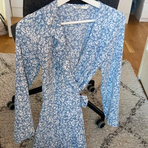 Blåvit blommig omlottklänning Only - Supersöt blå och vit omlottklänning från Only med blommigt mönster. Klänningen har långa ärmar, knytband i midjan och v-ringad hals. Perfekt för vår och sommar när du vill känna dig extra fin.