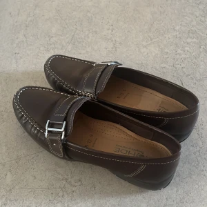 Bruna loafers med spänne - Snygga bruna loafers i skinn med dekorativt metallspänne och kontrastsömmar. Klassisk design med rund tå och platt sula, perfekta för en stilren look. Bekväma och tidlösa skor som passar till många outfits.