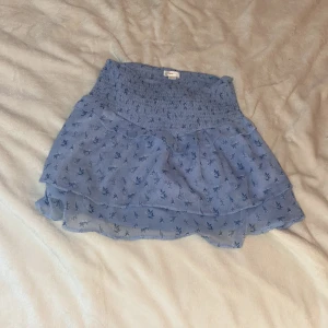 Blommig blå volangkjol Gina Tricot - Supersöt blå kortkjol från Gina Tricot med små blommor och volangdetaljer. Kjolen har smock i midjan för en bekväm passform och är gjord i ett lätt, luftigt material 