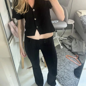 Svarta bootcut-byxor från Gina Tricot - Säljer ett par svarta bootcut-byxor från Gina Tricot med låg midja och två knappar framtill. Byxorna har pressveck längs benen och är i ett stretchigt material som sitter snyggt. Perfekta om du gillar en stilren och trendig look.