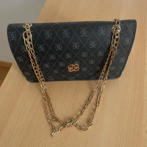 Svart axelväska med kedja från By Malene Birger - Snygg svart axelväska från By Malene Birger med guldiga kedjor och klassiskt monogrammönster över hela väskan. Väskan har ett elegant guldemblem framtill och är tillverkad i tåligt syntetmaterial. Perfekt storlek för det viktigaste och ger en lyxig vibe till din outfit.